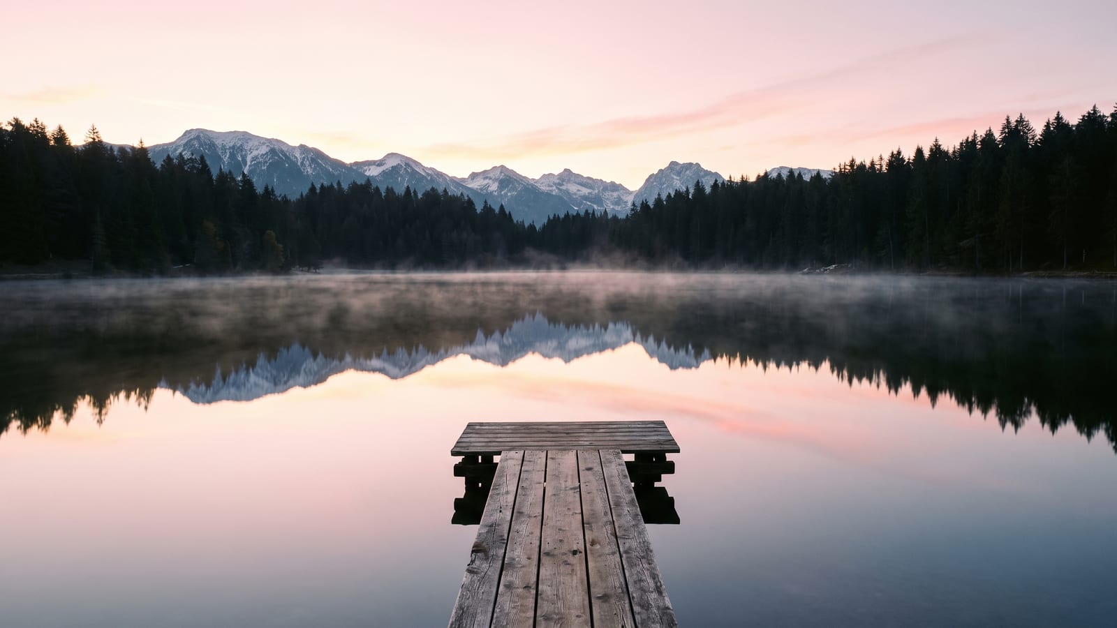A weathered wooden jetty reaching into a still alpine lake at dawn, with pine forest and snow-dusted peaks behind